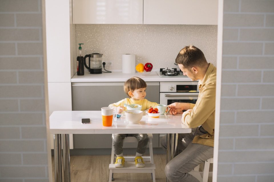 Litet barn på en hög stol med fotstöd äter vid köksbordet med sin pappa